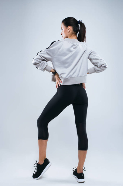 schlanke und sportliche junge Frau posiert und macht Sportübungen stehend mit dem Rücken zur Kamera im Studio vor grauem Hintergrund - Foto, Bild