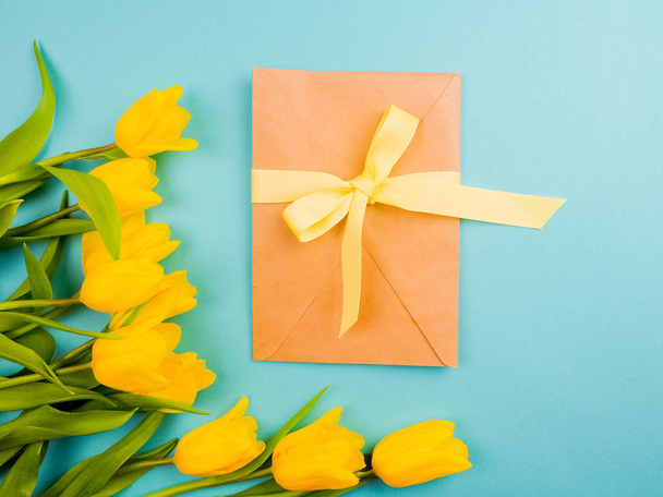 Любовь, романтика, поздравления, концепция праздника - желтые тюльпаны на голубом фоне. нежные лепестки тюльпана с подарочным конвертом в центре
 - Фото, изображение