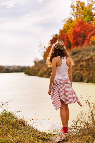 autumn, harmony, travelling concept. young girl dressed in sportswear standing on the river bank, on the background there is forest of bright red and yellow trees like in fire - Photo, Image