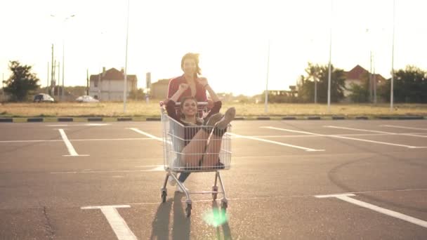 Mujer joven está sentada en el carrito de la compra, mientras su amiga la empuja hacia atrás en el aparcamiento junto al centro comercial, disfrutando al aire libre con la carrera de carrito de la compra durante el atardecer. Lente llamarada
 - Metraje, vídeo