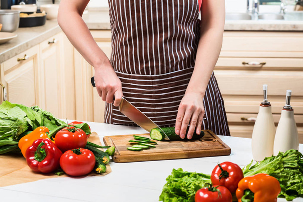 Mladá žena vaření v kuchyni doma. Žena řezy okurky a zeleninu s nožem. - Fotografie, Obrázek