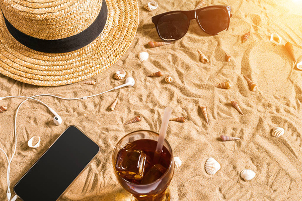 Ropa de playa de verano, gafas de sol, sombrero, bebida fría en un vaso y conchas marinas en la playa de arena
. - Foto, Imagen