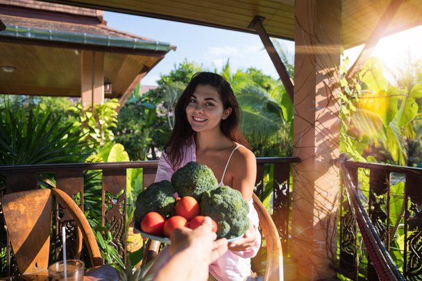 Männliche Hand gibt schöne Frau Teller mit frischem Gemüse pov Mädchen glücklich lächelnd auf Sommerterrasse im tropischen Wald sitzen - Foto, Bild