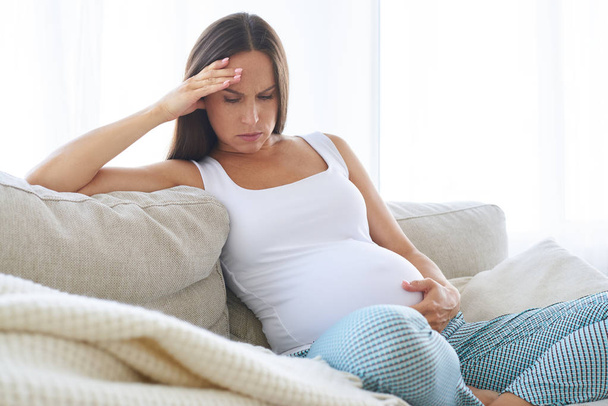 Disappointed pregnant woman sitting on sofa with hand on belly  - Fotografie, Obrázek