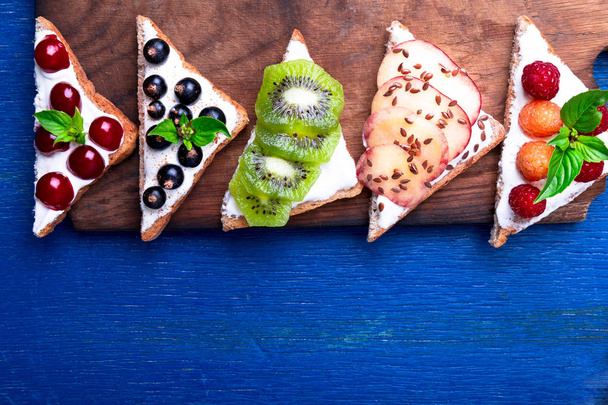 Fruit toast on wooden board on blue rustic background. Healthy breakfast. Clean eating. Dieting concept. Grain bread slices with cream cheese and various fruit, berries, seeds. Vegetarian. Top view. - 写真・画像