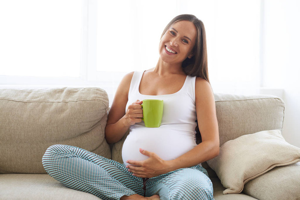Pregnant woman sitting with cup of tea on sofa  - Photo, Image