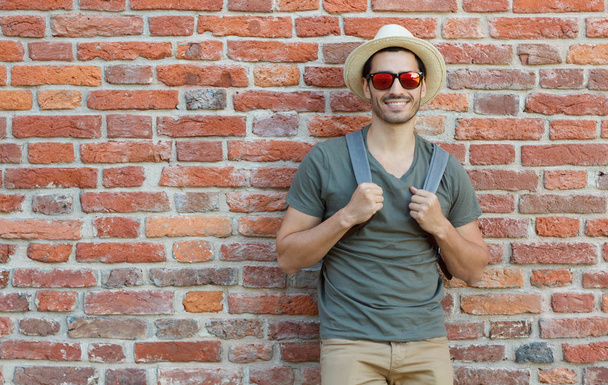 Outdoor portrait of young Caucasian man pictured with red brick wall in background looking through sunglasses with joyful smile, pulling straps of backpack forward as if ready to continue journey - 写真・画像
