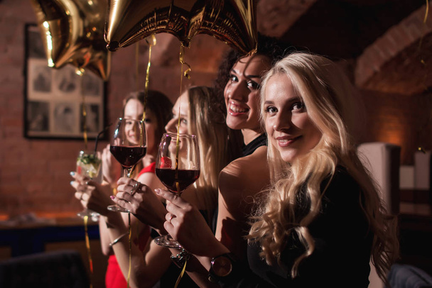 Χαρούμενα νεαρές γυναίκες στέκεται κοιτάζοντας πίσω κάμερα κρατώντας ποτήρια κρασί γιορτάζει τα γενέθλια της φίλης τους στο εστιατόριο - Φωτογραφία, εικόνα