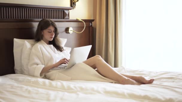 Woman in a bathrobe using her laptop in bed - Кадри, відео