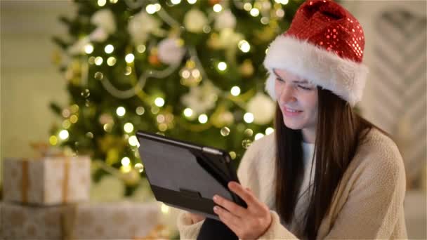 Closeup Portrait of Cute Brunette with Tablet in Her Hands on Christmas Tree Background. View of Emotional Girl in Santa Hat Smiling and Using Gadget During the Celebration of Xmas. - Séquence, vidéo