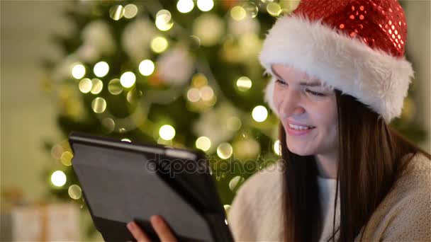 Emotional Woman in Santa Hat Using Gadget During the Celebration of Xmas. Amazing Brunette with Long Hair and Charming Smile Touching a Screen of Her Tablet at Christmas Decorated Home. - Filmati, video