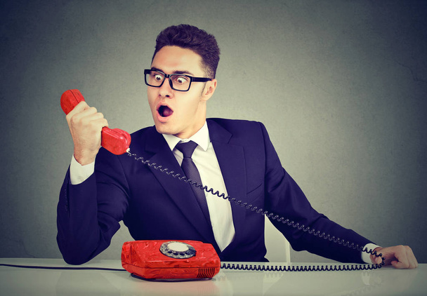 Shocked business man receiving bad news on the phone sitting at desk in his office  - 写真・画像