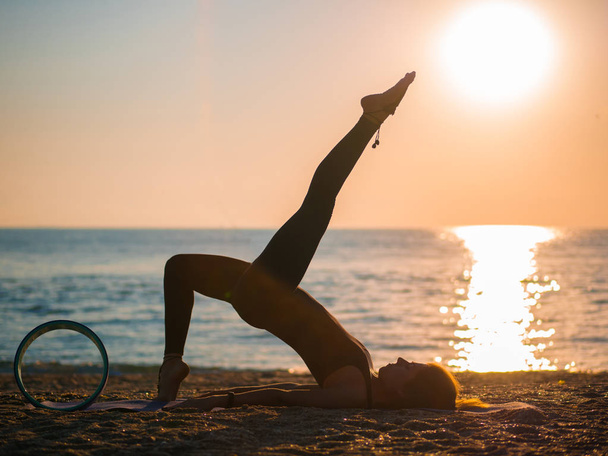 Silhouette of beautiful young woman practicing morning yoga on sand sea beach with yoga wheel. - 写真・画像