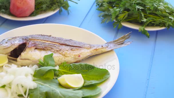 Grilled sea bream on plate with lemon, arugula, slice of lemon and onions. Healthy food. Blue table background. - Footage, Video