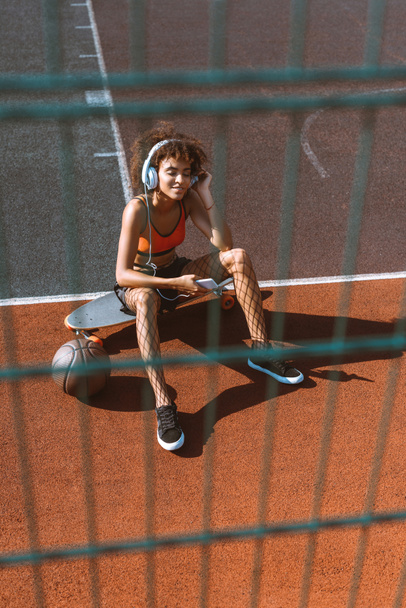 junge afrikanisch-amerikanische Frau in Sportbekleidung hört Musik über Kopfhörer und sitzt auf dem Longboard am Sportplatz - Foto, Bild