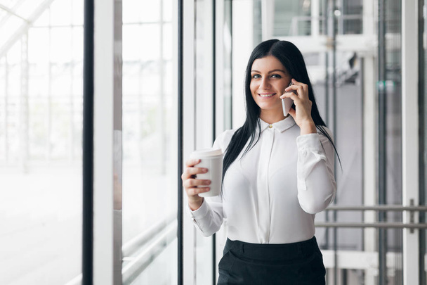 Successful business woman with coffee and smartphone in an office setting - Foto, afbeelding