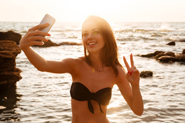 Joyful brunette woman in bikini making selfie on her smartphone - Foto, imagen