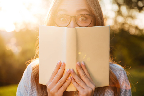 Young brunette woman in eyeglasses hiding behind a book - Fotó, kép