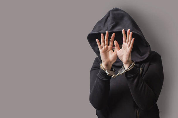 a girl in a black hood with handcuffed hands hides her face. isolated on a gray background. - Фото, зображення
