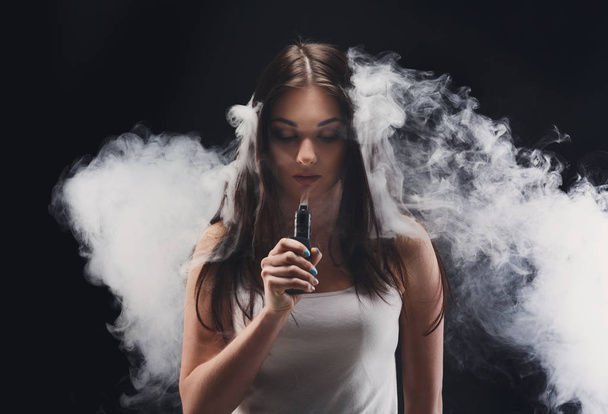 Young woman vaping e-cigarette with smoke on black - Zdjęcie, obraz