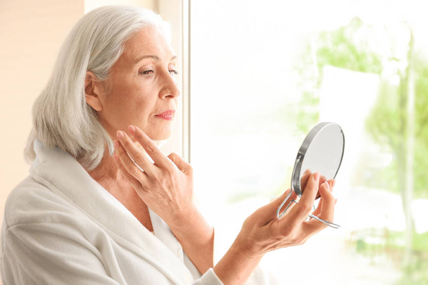Bella donna anziana che tiene lo specchio e applica la crema per il viso a casa
 - Foto, immagini