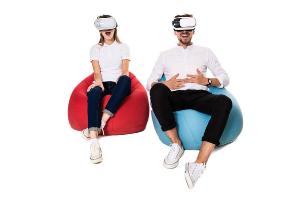 Emocionada pareja joven experimentando realidad virtual sentada en bolsas de frijoles aisladas sobre fondo blanco
 - Foto, Imagen