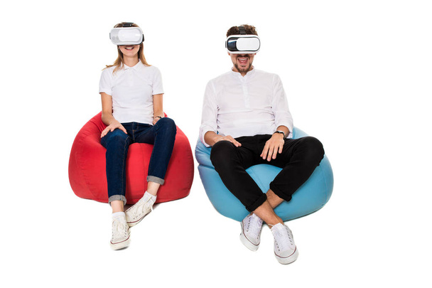 Возбужденная молодая пара испытывает виртуальную реальность, сидя на бобовых мешках изолированы на белом фоне
 - Фото, изображение