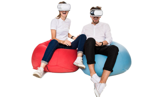 Возбужденная молодая пара испытывает виртуальную реальность, сидя на бобовых мешках изолированы на белом фоне
 - Фото, изображение