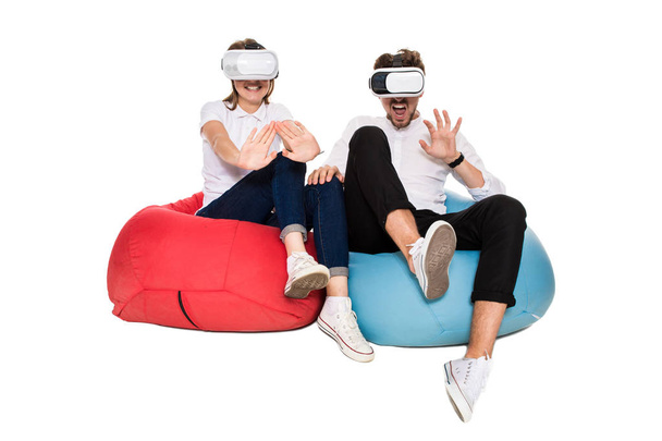 Excited young couple experiencing virtual reality seated on beanbags isolated on white background - Фото, зображення