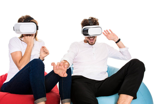 Excited young couple experiencing virtual reality seated on beanbags isolated on white background - Foto, Imagem
