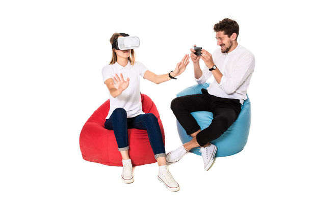 Smiling friends experiencing virtual reality glasses seated on beanbags isolated on white background. - Photo, Image