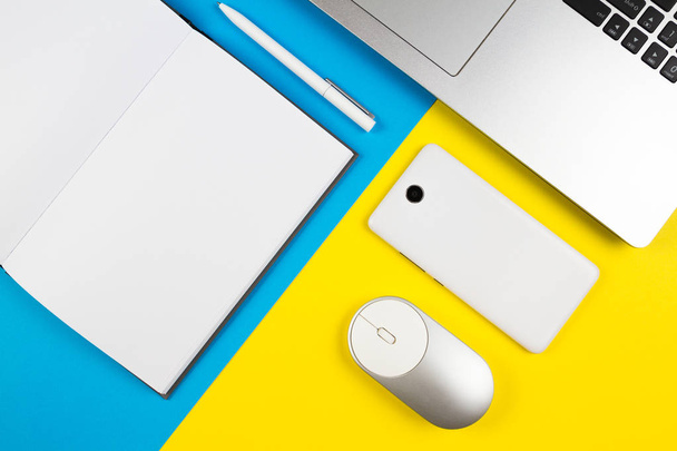 Modern workplace with notebook, computer mouse, mobile phone and white pen on blue and yellow color background - Photo, Image