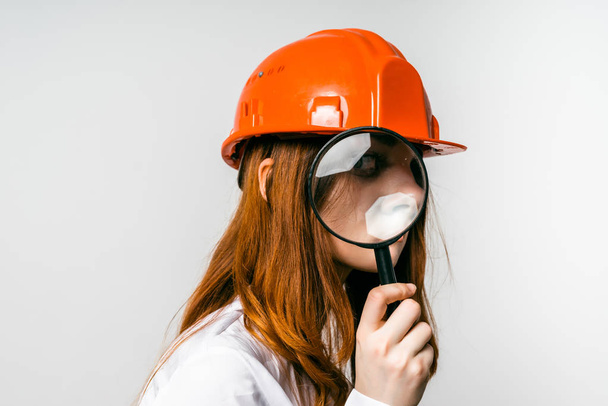 girl with a magnifying glass in her hand in a construction helmet - Photo, Image