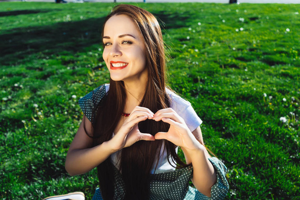 Beautiful woman shows heart shape with hands,sit on grass - Zdjęcie, obraz
