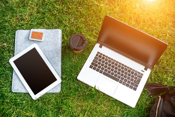 Комп'ютер ноутбука на зеленій траві з чашкою кави, сумкою та планшетом у відкритому парку
 - Фото, зображення