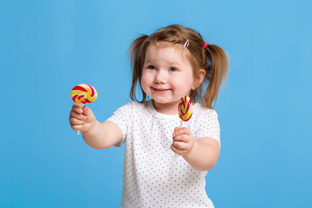 Prachtige vrouwelijke kindje houden grote lolly spiraal snoep glimlachend gelukkig geïsoleerd op blauwe achtergrond. - Foto, afbeelding