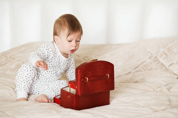 Κοριτσάκι θαυμάζει τα αξεσουάρ στο κουτί. Κοριτσάκι παίζει με τα κοσμήματα της μητέρας κάθεται σε ένα κρεβάτι σε πιτζάμες ξυπόλυτος - Φωτογραφία, εικόνα