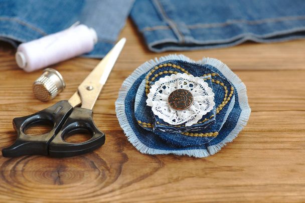 ハンドメイドデニムフラワージュエリー。はさみ、糸、シンブル、針、木の背景に女性の古いジーンズ。古いジーンズをリサイクルするための縫製プロジェクト。引き裂かれたジーンズをどうするか。閉鎖 - 写真・画像