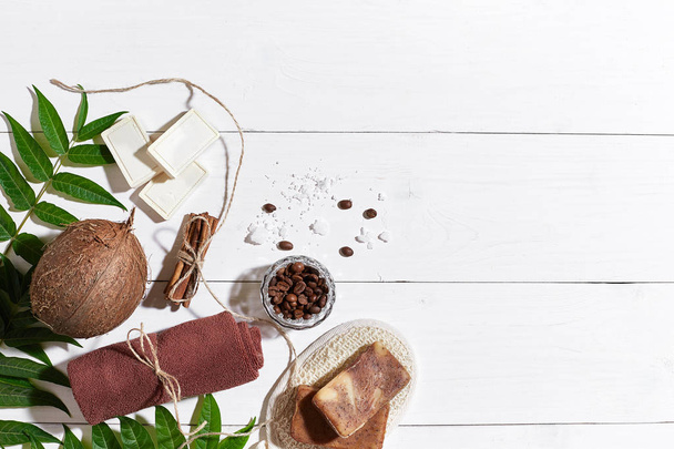 natürliche handgemachte Seifen mit Kaffeebohnen, Zimt, Meersalz, Luffa, braunem Handtuch, Kokosnuss und grünen Blättern auf weißem Holzgrund - Foto, Bild