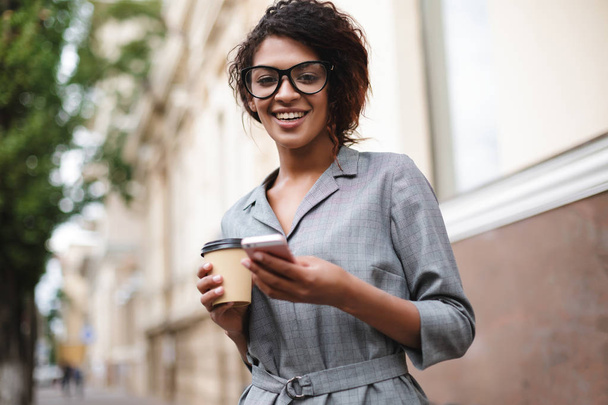 Χαμογελώντας αφρικανικό αμερικανικό κορίτσι με τα γυαλιά που στέκεται στο δρόμο με κινητό τηλέφωνο και καφέ στα χέρια ενώ ευτυχώς ψάχνει στην κάμερα - Φωτογραφία, εικόνα