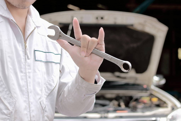 Portrait of car mechanic man holding wrench and her hand with i love you sign with car in open hood at the repair garage background. - Photo, Image