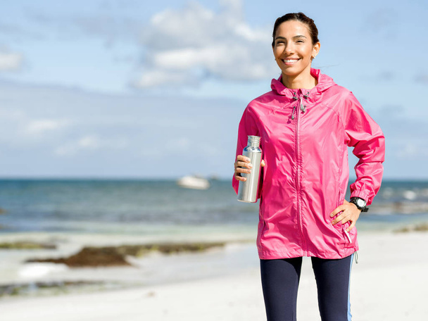 Beautiful girl in sport clothes drinking water after workout on the beach - Fotoğraf, Görsel