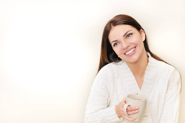 A happy woman with a cup of tea wears a white sweater smiling - 写真・画像