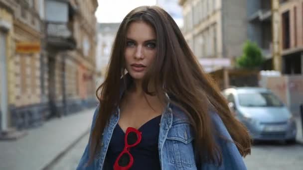 Brunette woman with long hair smiles and whirls walking along the street in her jeans jacket - Кадри, відео