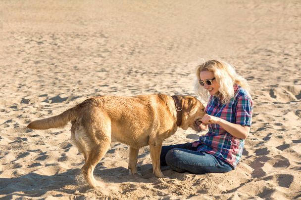 Portrait of young beautiful woman in sunglasses sitting on sand beach with golden retriever dog. Girl with dog by sea. - Photo, Image