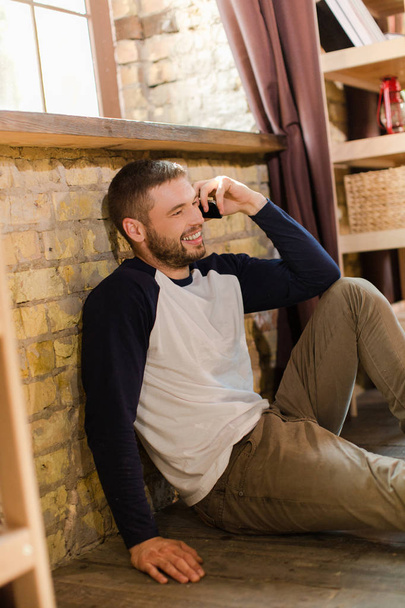 Όμορφος άντρας κάθεται στο πάτωμα δίπλα στο παράθυρο μιλώντας στο τηλέφωνο. - Φωτογραφία, εικόνα