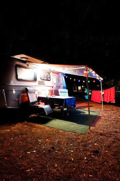 Retro vintage trailer caravana de acampamento à noite em uma floresta com luzes, cadeiras e uma mesa
 - Foto, Imagem