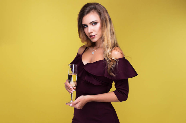 Sexy blond młoda kobieta trzyma kieliszek szampana, patrząc na kamery i uśmiech. Atrakcyjne kobiety w sexy sukienka w studio na żółtym tle - Zdjęcie, obraz
