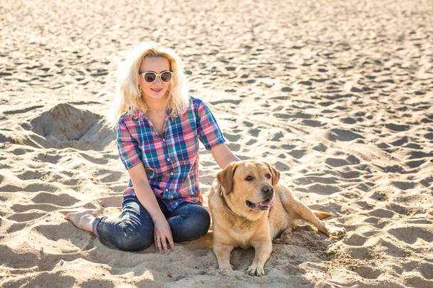 Portret van jonge mooie vrouw in zonnebril zittend op zand strand met gouden retriever hond. Meisje met hond aan zee. - Foto, afbeelding