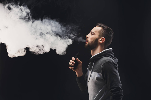 Young man vaping e-cigarette with smoke on black - Foto, Bild